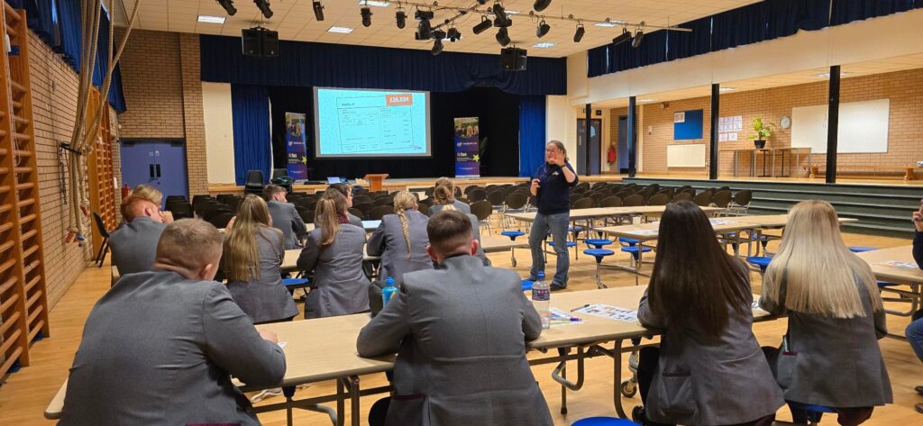 A school hall with pupils sat on benches learning financial education from a MyBnk trainer talking in front of a whiteboard screen