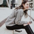 A smiling university student sat outside in a city with a phone in her hand and wallet next to her