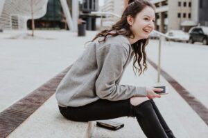 A smiling university student sat outside in a city with a phone in her hand and wallet next to her