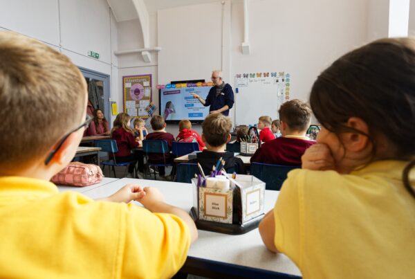 Primary school children in a classroom looking at the teacher standing at a digital whiteboard