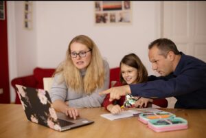 wo adults and a child sat around a dining table looking at a laptop together