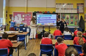 Primary school children in a classroom looking at two teachers standing at a digital whiteboard